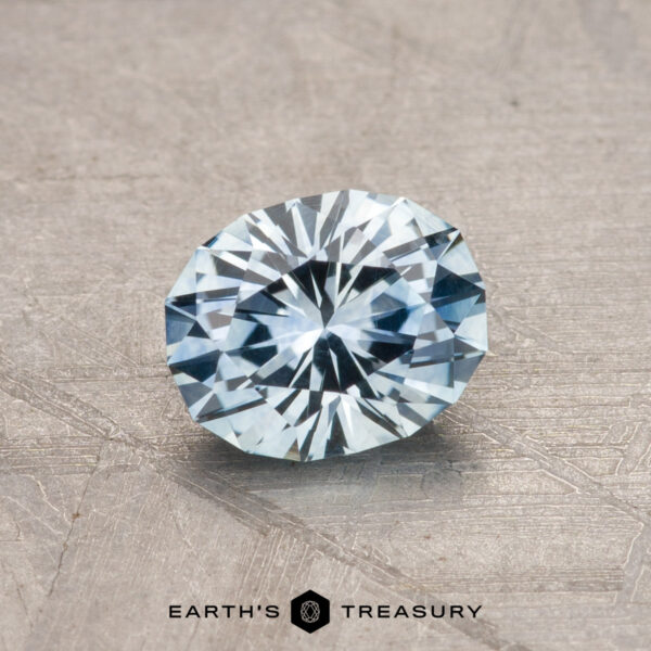 1.05-Carat Montana Sapphire (Heated) A 1.05-carat Montana Sapphire (heated) with a faceted, oval cut and light blue hue is placed on a textured surface.