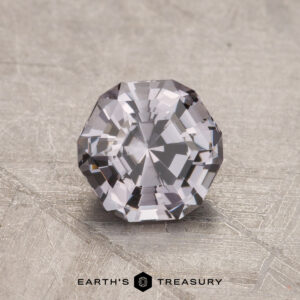A faceted, clear 1.11-carat Spinel with an octagonal shape rests on a textured surface.