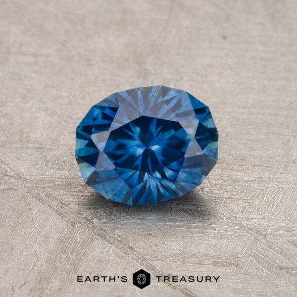 0.91-Carat Montana Sapphire (Heated) A 0.91-carat Montana Sapphire (heated), featuring a round faceted cut and blue hue, is showcased on a textured gray surface with the text "Earth's Treasury" below.