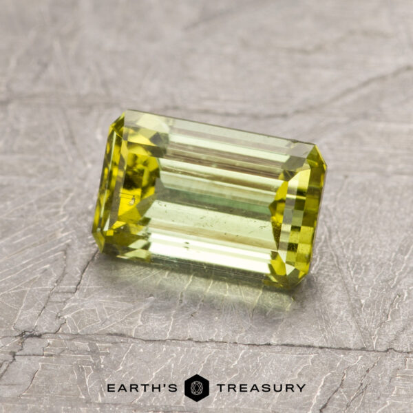 1.18-Carat Greenish Yellow California Tourmaline The ET1443 gemstone, featuring a faceted cut, sits on a textured gray surface in a rectangular green form.