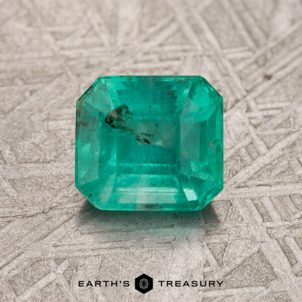 1.14-Carat Bright Green Colombian Emerald (Oiled) A treated 1.14-carat Colombian emerald with a rich green hue, cut in an emerald shape, rests on a textured surface, featuring the words "Earth's Treasury" below it.