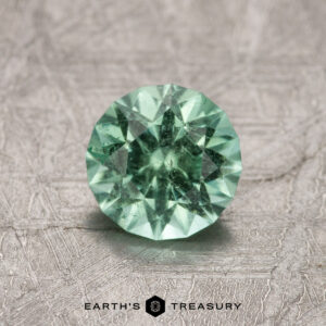 A 1.02-carat Maine Tourmaline featuring a round, faceted mint green design is displayed on a textured surface.