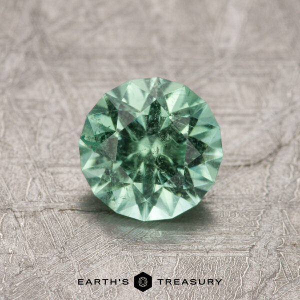 A 1.02-carat Maine Tourmaline featuring a round, faceted mint green design is displayed on a textured surface.