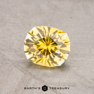 A yellow faceted gemstone on a textured surface with the logo "Earth's Treasury" below, showcasing the 1.15-Carat Montana Sapphire (Heated).