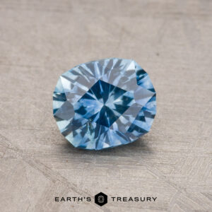 A sparkling blue 1.07-carat Montana sapphire (heated) displayed on a textured surface with the text "Earth's Treasury" below.