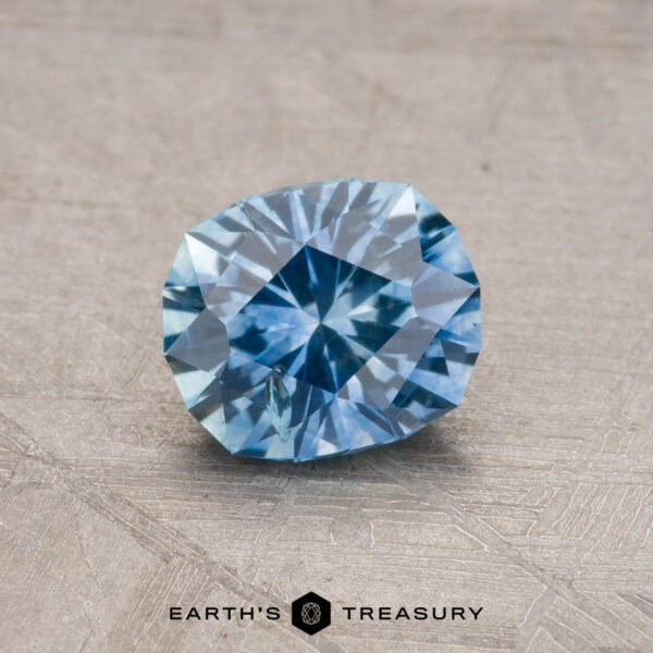 A sparkling blue 1.07-carat Montana sapphire (heated) displayed on a textured surface with the text "Earth's Treasury" below.