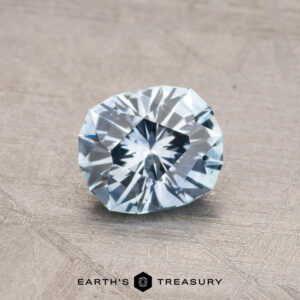 A faceted, pale blue gemstone is displayed against a textured gray background with "Earth's Treasury" written at the bottom, showcasing the 0.99-Carat Montana Sapphire (Heated).