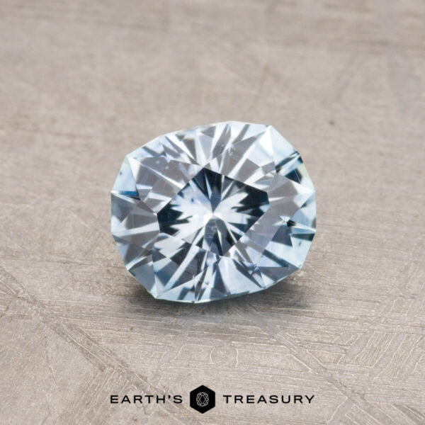 0.99-Carat Montana Sapphire (Heated) A faceted, pale blue gemstone is displayed against a textured gray background with "Earth's Treasury" written at the bottom, showcasing the 0.99-Carat Montana Sapphire (Heated).