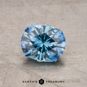 A 1.23-carat Montana sapphire (heated) with intricate cuts is displayed on a textured surface, showcasing its clarity and brilliance. The "Earth's Treasury" logo is visible at the bottom.