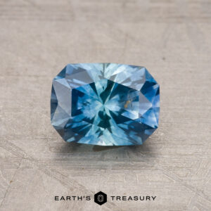 A faceted blue 1.24-carat Montana sapphire (heated) on a textured surface with the logo "Earth's Treasury" below it.