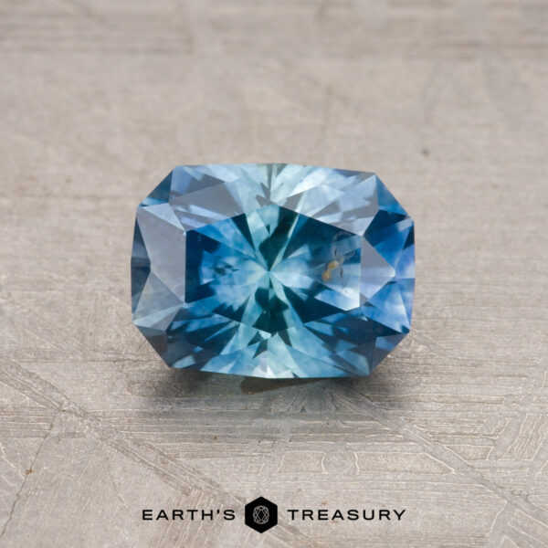 1.24-Carat Montana Sapphire (Heated) A faceted blue 1.24-carat Montana sapphire (heated) on a textured surface with the logo "Earth's Treasury" below it.