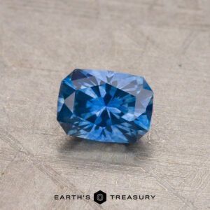 A 0.90-carat Montana Sapphire, heated for enhanced color, sits on a textured surface with the "Earth's Treasury" logo at the bottom.