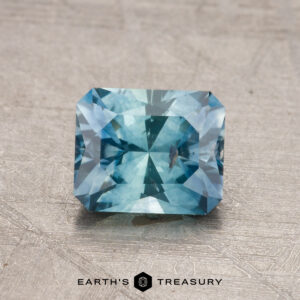 A 1.28-carat heated Montana sapphire with a radiant cut and intricate facets, displayed on a textured surface.