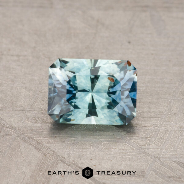 1.06-Carat Montana Sapphire (Heated) A rectangular, blue-green 1.06-carat Montana sapphire with facets rests on a textured gray surface. Logo text at the bottom reads: "Earth's Treasury.