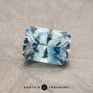 A 1.00-carat Montana sapphire (heated), featuring a rectangular, faceted design with a reflective surface, is elegantly displayed on a textured gray background.