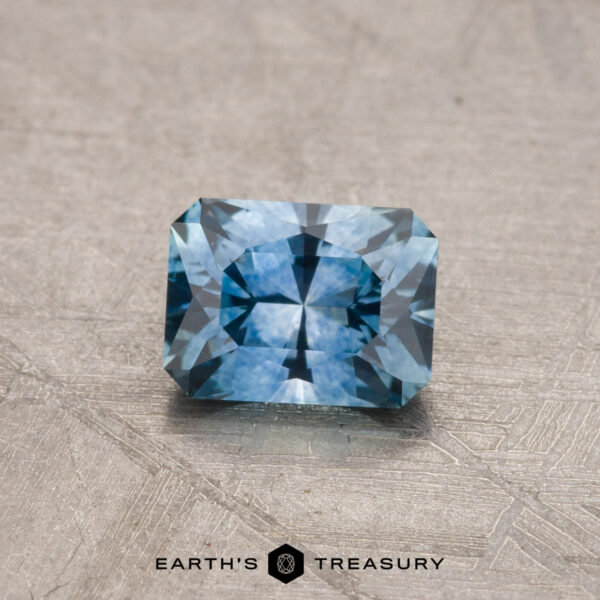 0.81-Carat Montana Sapphire (Heated) A rectangular faceted-cut blue gemstone, identified as a 0.81-carat Montana Sapphire (Heated), is showcased on a textured surface with the Earth's Treasury logo visible below.