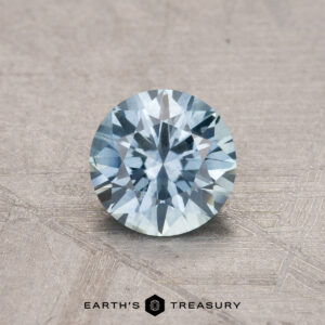 Displayed on a textured surface, this round, faceted blue gemstone is labeled with the "Earth's Treasury" logo and identified as a 0.90-carat Montana Sapphire (heated).