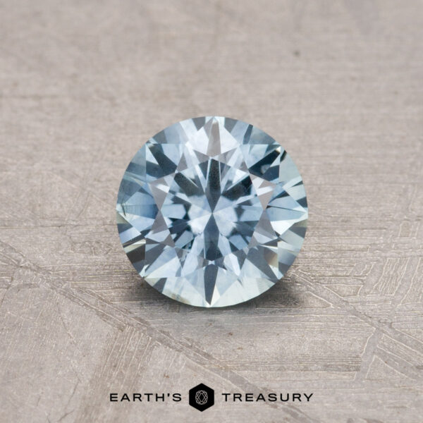 0.90-Carat Montana Sapphire (Heated) Displayed on a textured surface, this round, faceted blue gemstone is labeled with the "Earth's Treasury" logo and identified as a 0.90-carat Montana Sapphire (heated).