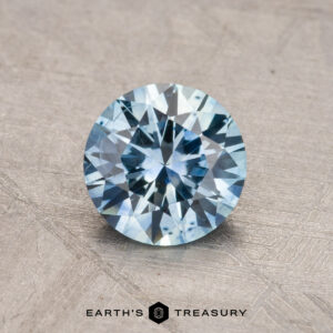 A 1.18-carat, round-cut Montana Sapphire (heated) in a light blue hue is displayed on a textured surface, with the logo "Earth's Treasury" beneath it.