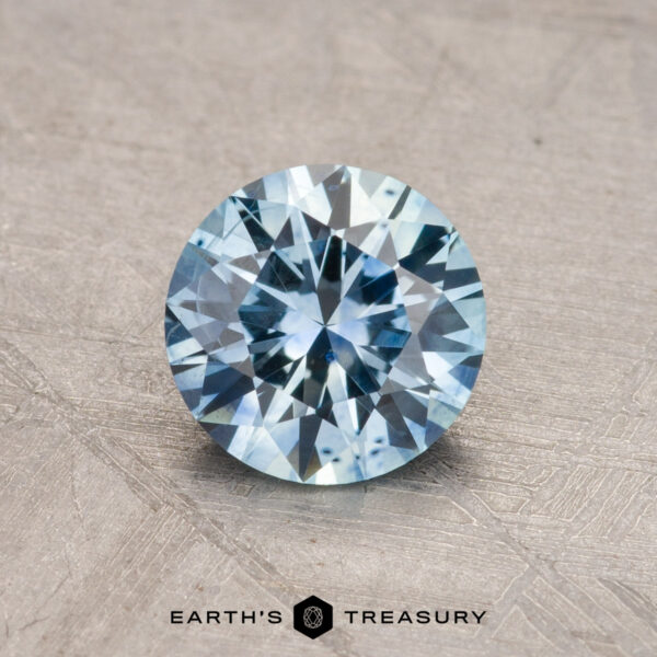 1.18-Carat Montana Sapphire (Heated) A 1.18-carat, round-cut Montana Sapphire (heated) in a light blue hue is displayed on a textured surface, with the logo "Earth's Treasury" beneath it.