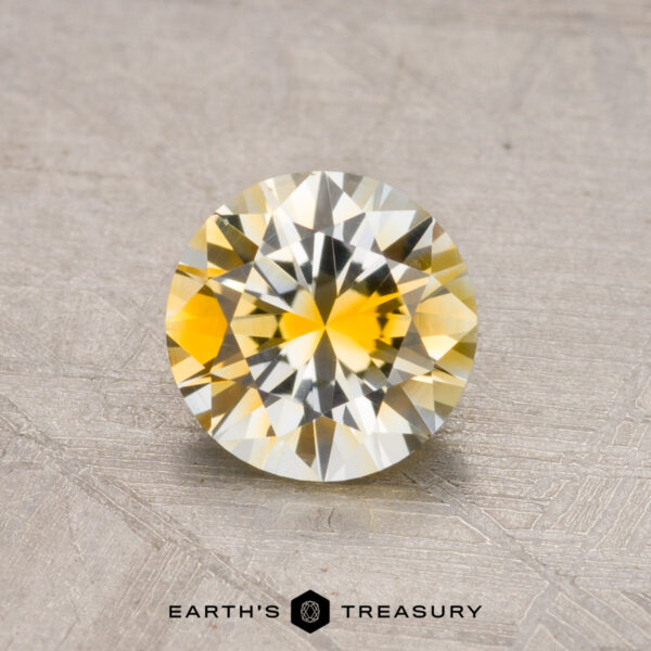 1.08-Carat Montana Sapphire (Heated) Faceted round yellow gemstone on a textured surface with the text "Earth's Treasury" at the bottom. Product: 1.08-Carat Montana Sapphire (Heated).