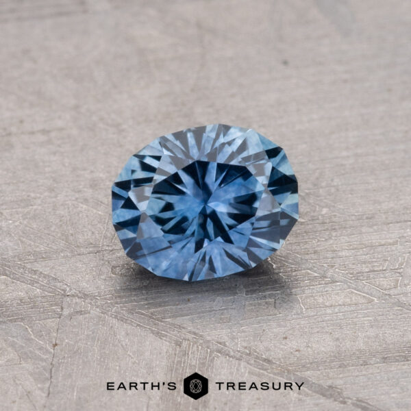 0.81-Carat Montana Sapphire (Heated) A round, faceted blue 0.81-carat Montana Sapphire (heated) placed on a textured gray surface, with the text "Earth's Treasury" at the bottom.