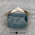 A textured blue rock elegantly sits on a flat surface, adorned with a gold triangular frame. Below it, the words "Earth's Treasury" gleam, evoking the timeless beauty of The "Alora" band in 14k yellow gold.