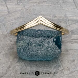 A textured blue rock elegantly sits on a flat surface, adorned with a gold triangular frame. Below it, the words "Earth's Treasury" gleam, evoking the timeless beauty of The "Alora" band in 14k yellow gold.