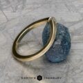 The "Alora" band in 14k yellow gold is gracefully perched on a textured blue stone, with a gray geometric pattern in the background framing the text "Earth's Treasury" at the bottom.