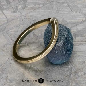 The "Alora" band in 14k yellow gold is gracefully perched on a textured blue stone, with a gray geometric pattern in the background framing the text "Earth's Treasury" at the bottom.