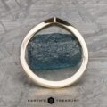 A 14k yellow gold "Alora" band gracefully wraps around a textured, cylindrical blue gemstone. The background showcases a patterned gray surface with the "Earth's Treasury" logo at the bottom, reflecting Alora's harmonious fusion of nature and art.
