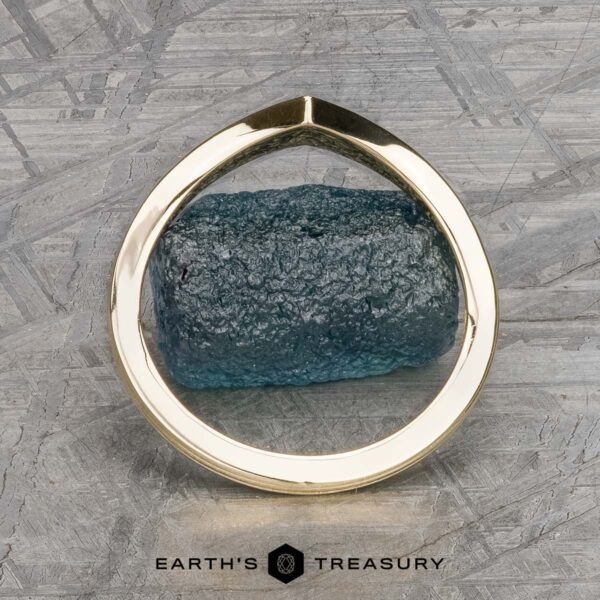 The "Alora" band in 14k yellow gold A 14k yellow gold "Alora" band gracefully wraps around a textured, cylindrical blue gemstone. The background showcases a patterned gray surface with the "Earth's Treasury" logo at the bottom, reflecting Alora's harmonious fusion of nature and art.