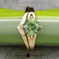 The "Alora" ring, crafted in 14k yellow gold and adorned with a 1.93-carat Australian gemstone, rests on a green and silver textured surface, reminiscent of the vibrant harmony created by the notes of an Alora Band melody.