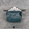 The "Aurelia" band in 14k white gold, featuring seven diamonds, rests atop a rough blue gemstone on a textured gray background. "EARTH'S TREASURY" appears at the bottom.