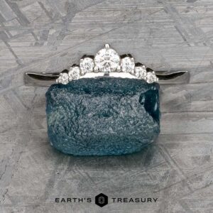 The "Aurelia" band in 14k white gold, featuring seven diamonds, rests atop a rough blue gemstone on a textured gray background. "EARTH'S TREASURY" appears at the bottom.