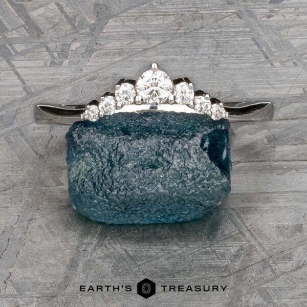 The "Aurelia" band in 14k white gold The "Aurelia" band in 14k white gold, featuring seven diamonds, rests atop a rough blue gemstone on a textured gray background. "EARTH'S TREASURY" appears at the bottom.