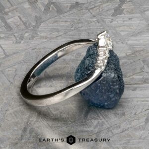 The "Aurelia" band in 14k white gold, featuring a row of baguette-cut gemstones, is displayed upright with its band resting on a rough blue stone against a textured gray background.