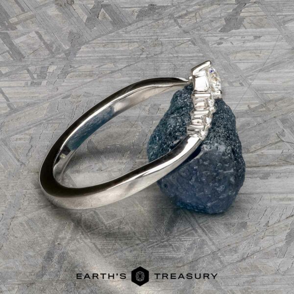 The "Aurelia" band in 14k white gold The "Aurelia" band in 14k white gold, featuring a row of baguette-cut gemstones, is displayed upright with its band resting on a rough blue stone against a textured gray background.