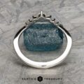 The "Aurelia" band in 14k white gold, featuring a jagged-edge setting with a rough blue gemstone, stands upright on a textured gray surface. The Earth's Treasury logo and Aurelia branding are visible at the bottom.