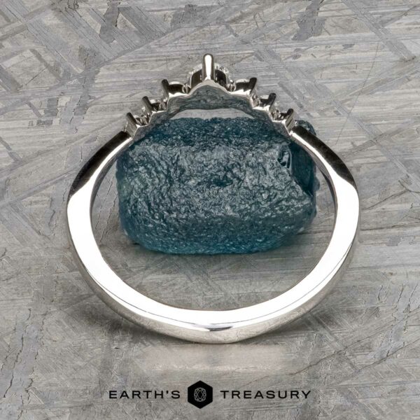 The "Aurelia" band in 14k white gold The "Aurelia" band in 14k white gold, featuring a jagged-edge setting with a rough blue gemstone, stands upright on a textured gray surface. The Earth's Treasury logo and Aurelia branding are visible at the bottom.