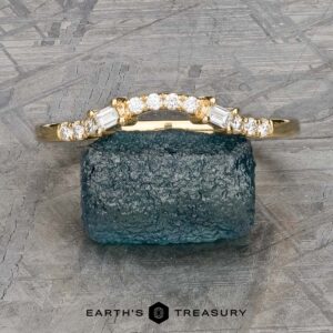 The "Fiona" band, crafted in 18k yellow gold and embellished with a series of small diamonds, is elegantly showcased on a rough greenish-blue stone. The backdrop presents a textured surface that amplifies the ring's captivating charm.