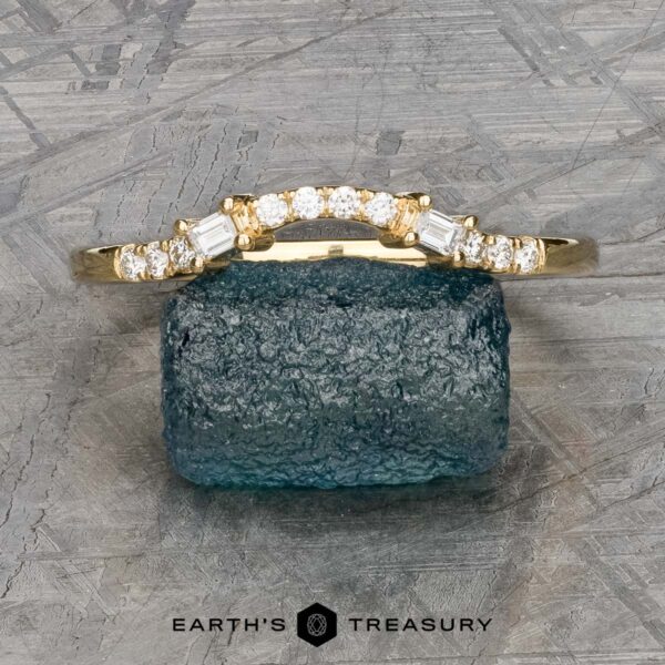 The "Fiona" band in 18k yellow gold The "Fiona" band, crafted in 18k yellow gold and embellished with a series of small diamonds, is elegantly showcased on a rough greenish-blue stone. The backdrop presents a textured surface that amplifies the ring's captivating charm.