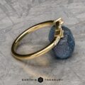 The "Fiona" band, crafted in 18k yellow gold and embellished with embedded diamonds, gracefully rests against a textured blue gemstone. Its allure is further enhanced by the sleek metallic surface in the background.