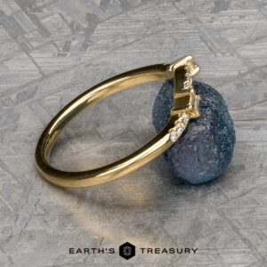 The "Fiona" band, crafted in 18k yellow gold and embellished with embedded diamonds, gracefully rests against a textured blue gemstone. Its allure is further enhanced by the sleek metallic surface in the background.
