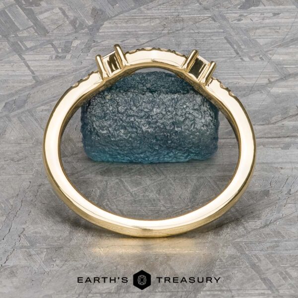 The "Fiona" band in 18k yellow gold The "Fiona" band in 18k yellow gold stands upright in front of a textured blue stone, with a logo reading "Earth's Treasury" at the bottom.