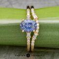 The "Fiona" ring, crafted in 18k yellow gold and featuring a stunning 1.80-carat Umba sapphire, is beautifully presented on a light green textured background, complemented by dainty round and rectangular clear stones.