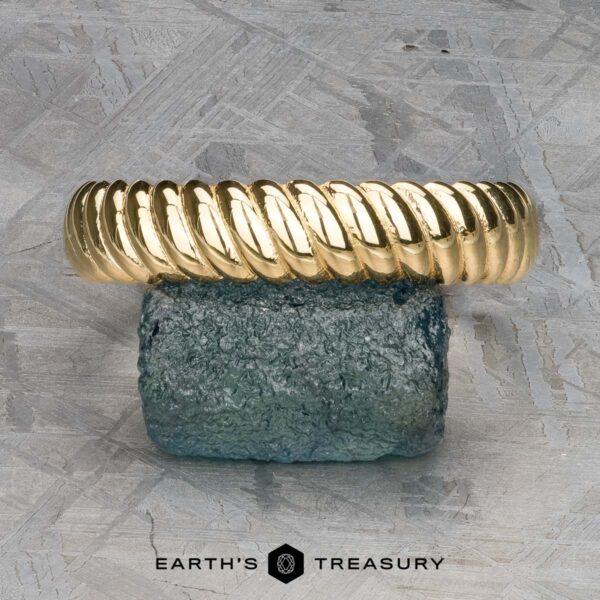 The "Damon" ring in 18k yellow gold The "Damon" ring in 18k yellow gold, a twisted masterpiece, elegantly rests on a textured blue stone with "Earth's Treasury" subtly inscribed below.