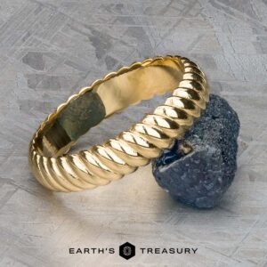 The "Damon" ring in 18k yellow gold, with its elegant twisted rope design, rests against a rugged gray stone on a textured surface. The text at the bottom reads "Earth's Treasury.