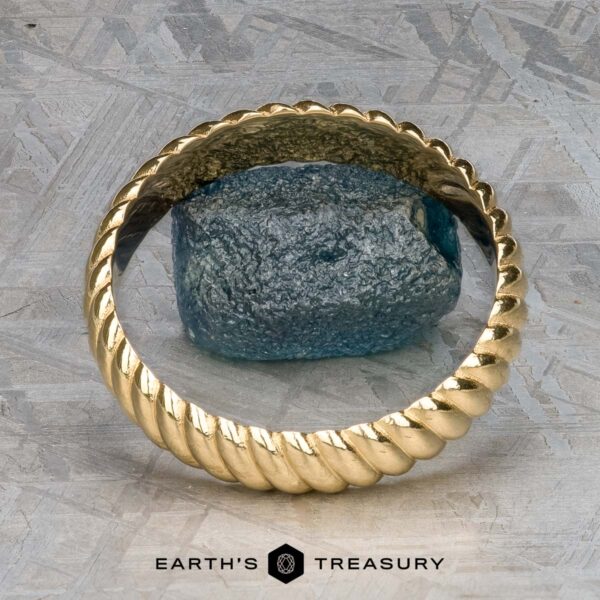 The "Damon" ring in 18k yellow gold An 18k yellow gold Damon ring elegantly encircles a raw blue gemstone, resting on a textured gray surface. Text below reads: "Earth's Treasury.