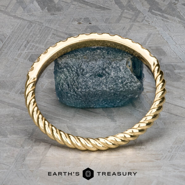 The "Lynn" band in 18k yellow gold The "Lynn" band in 18k yellow gold features a twisted design and textured finish, artfully balanced on a rugged blue stone with a metallic background. Text reads "Earth's Treasury.